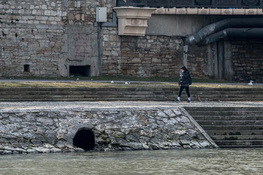 Фотографија од кејот на Вардар, на која се гледаат отвори за одливи во реката. Човек се движи по кејот.